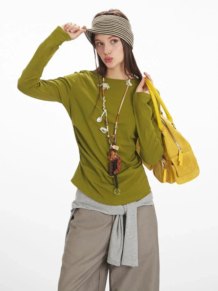 Model posing with a long-sleeve olive green top with ruched side detail, grey layered vest, taupe wide-leg pants, yellow handbag, and striped headband.