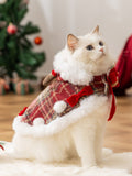 A white cat wearing a Christmas cape with a plaid pattern, faux fur trim, and small red bows, sitting in a festive setting.