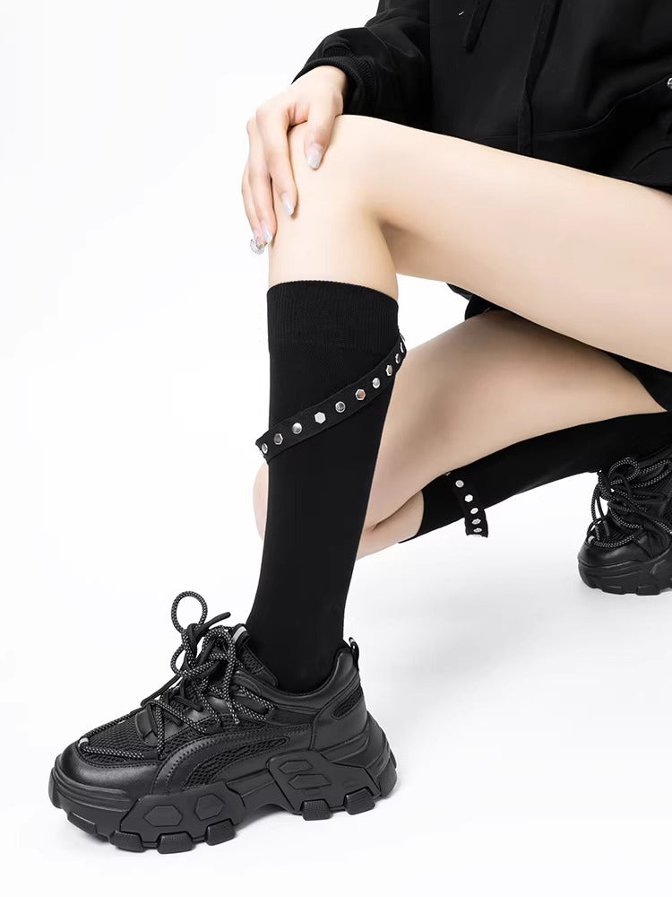 Person wearing black knee-high socks with stud accents and a strap, in a seated pose with chunky sneakers.