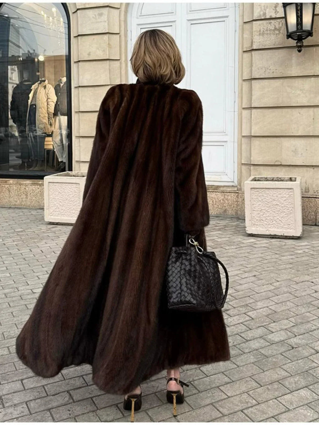 Rear view of a woman wearing a long brown mink fur coat, black pants, and brown heels walking on a cobblestone street.