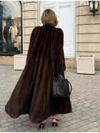 Rear view of a woman wearing a long brown mink fur coat, black pants, and brown heels walking on a cobblestone street.