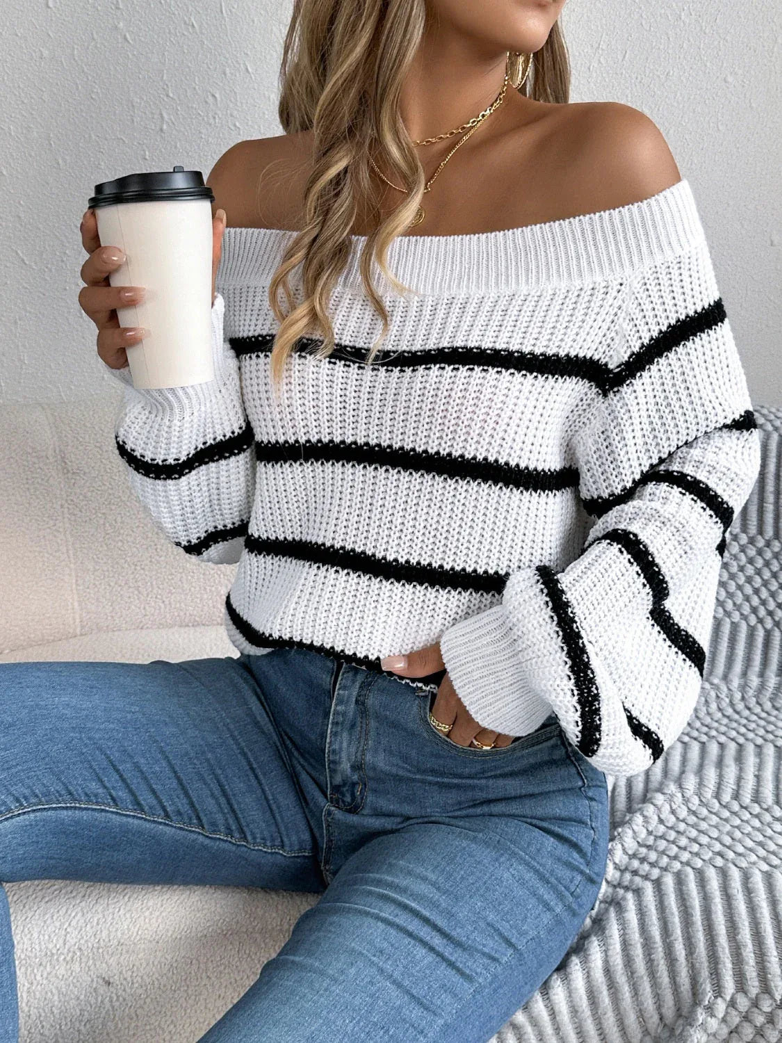 Woman wearing a white and black striped off-the-shoulder knit sweater with blue jeans, holding a coffee cup