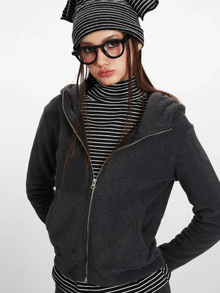 Woman in a charcoal grey zip-up hooded fleece jacket, striped turtleneck, and glasses, posing with hands in pockets.