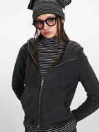 Woman in a charcoal grey zip-up hooded fleece jacket, striped turtleneck, and glasses, posing with hands in pockets.
