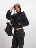 Woman adjusting her baseball cap while wearing a black cropped puffer jacket with a hood, grey t-shirt, and camouflage cargo pants, holding a black tote bag.