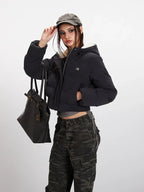 Woman adjusting her baseball cap while wearing a black cropped puffer jacket with a hood, grey t-shirt, and camouflage cargo pants, holding a black tote bag.