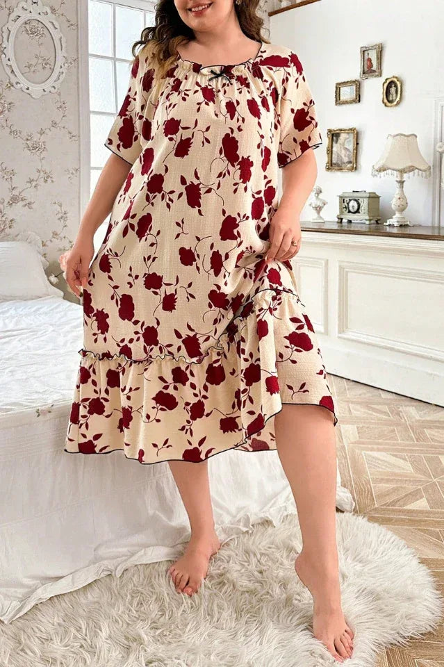 Plus-size woman wearing a cream-colored short-sleeve nightgown with a red floral print and ruffled hem, standing in a bedroom.
