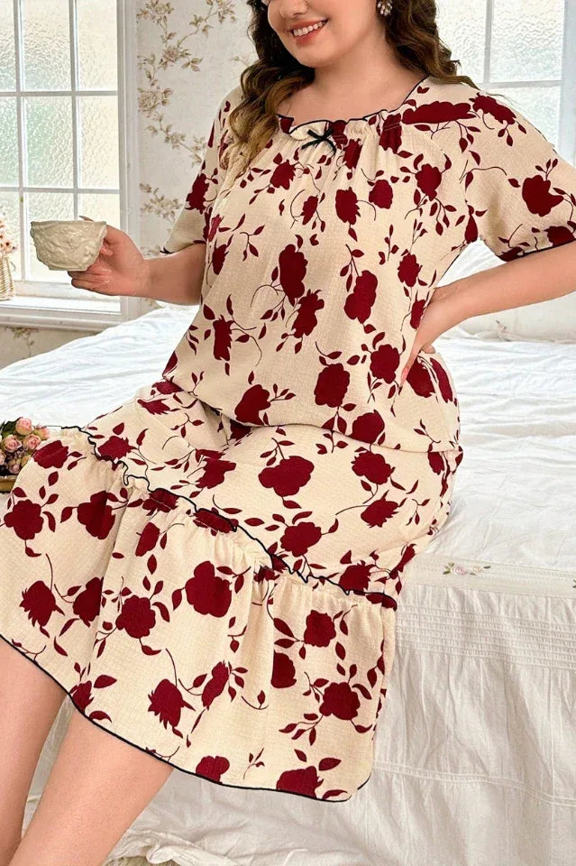 Plus-size woman wearing a cream-colored short-sleeve nightgown with a red floral print and ruffled hem, sitting on a bed and holding a teacup.