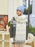 A woman stands looking to the side, wearing a light blue knit beanie and a white and blue striped knit scarf with fringe.