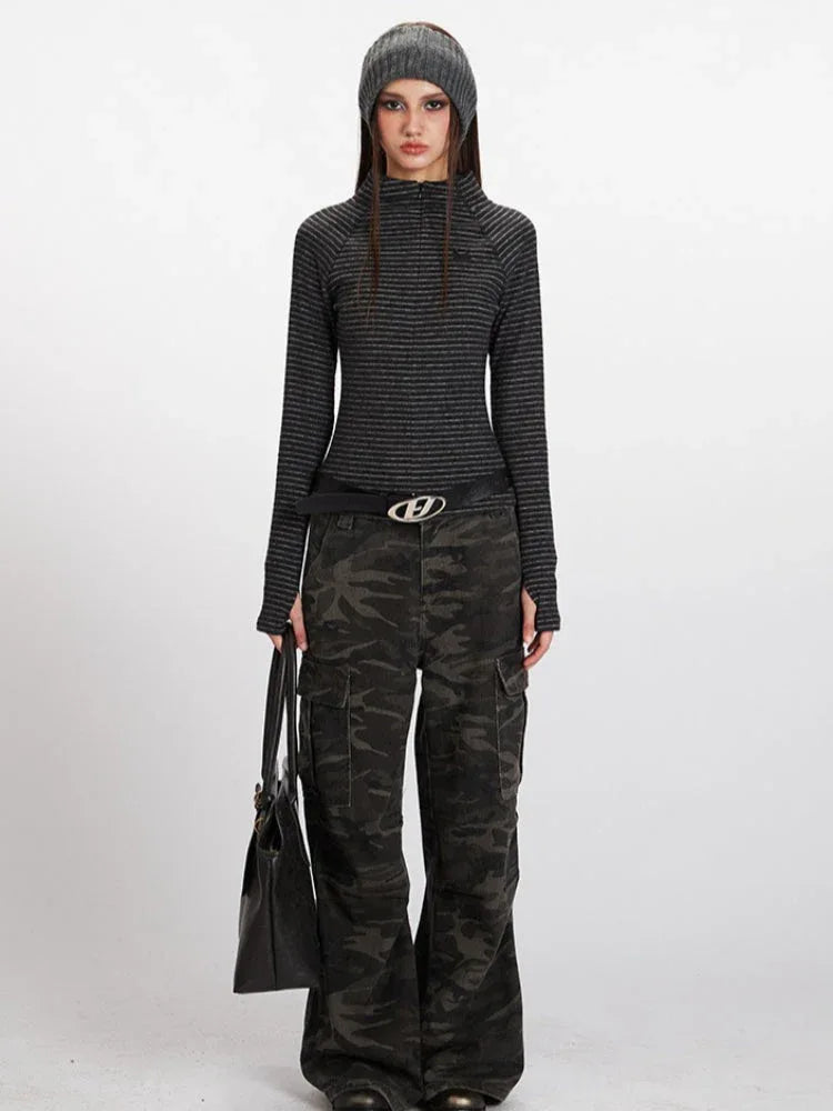 Woman wearing a grey striped long-sleeve mock neck top with a quarter zip closure and thumbholes, paired with camouflage cargo pants and a black beanie.