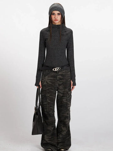 Woman wearing a grey striped long-sleeve mock neck top with a quarter zip closure and thumbholes, paired with camouflage cargo pants and a black beanie.