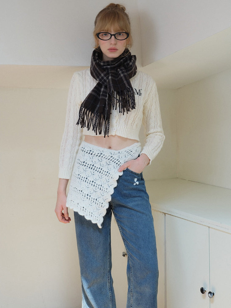 Woman wearing a cream cable knit cropped sweater with embroidered initial, a plaid scarf, and a white crocheted overlay skirt.