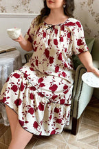 Plus-size woman wearing a cream-colored short-sleeve nightgown with a red floral print and ruffled hem, sitting in a chair and holding a teacup.