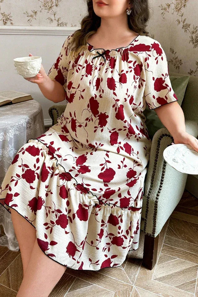 Plus-size woman wearing a cream-colored short-sleeve nightgown with a red floral print and ruffled hem, sitting in a chair and holding a teacup.