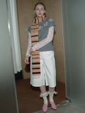 Woman wearing a grey short-sleeve sweater, white culottes, lace sleeves, a multi-colored striped scarf, and pink shoes standing with hands behind back.