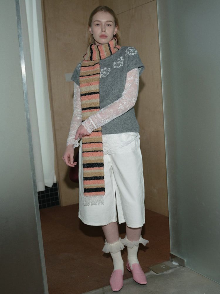 Woman wearing a grey short-sleeve sweater, white culottes, lace sleeves, a multi-colored striped scarf, and pink shoes standing with hands behind back.