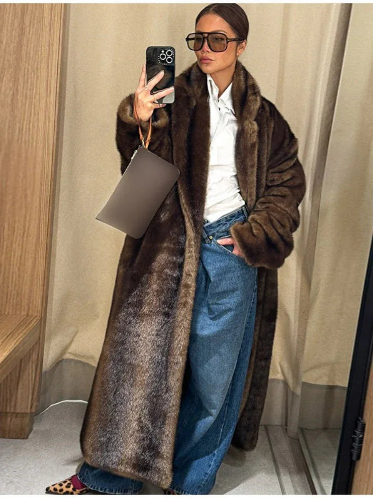 Woman wearing a long brown faux fur coat, blue jeans, and sunglasses, holding a clutch.