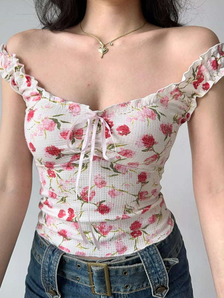 Close-up of a woman wearing a white floral print top with pink flowers and green leaves, featuring ruffled off-the-shoulder cap sleeves and a sweetheart neckline with a pink ribbon tie.