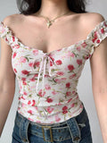 Close-up of a woman wearing a white floral print top with pink flowers and green leaves, featuring ruffled off-the-shoulder cap sleeves and a sweetheart neckline with a pink ribbon tie.
