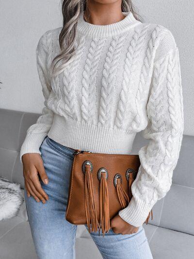 Woman holding a brown fringed clutch while wearing a white cropped cable knit sweater and light wash jeans