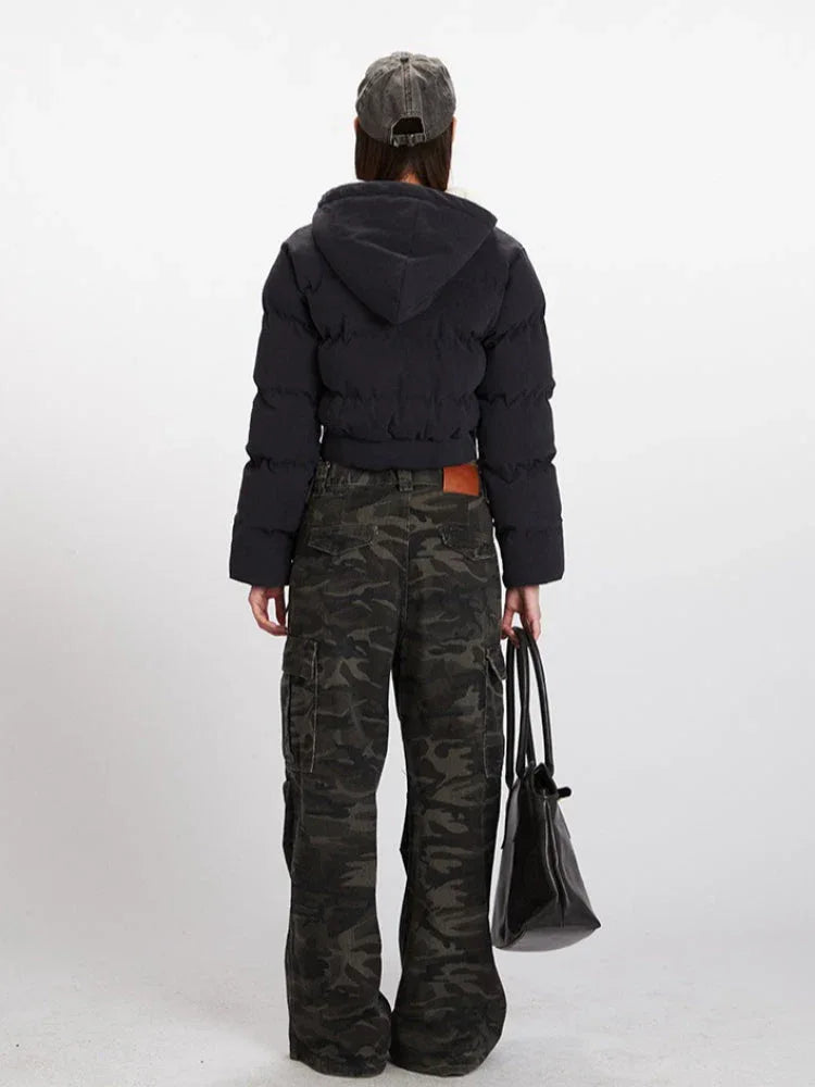 Back view of a woman wearing a black cropped puffer jacket with a hood, grey t-shirt, and camouflage cargo pants, holding a black tote bag.