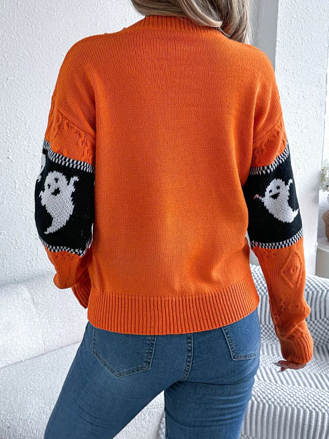 Woman wearing an orange crew neck knit sweater with a ghost print pattern on the sleeves and a black and white striped band across the chest.