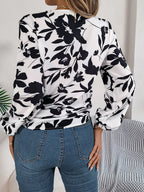 Back view of a woman wearing a white long-sleeve blouse with a black floral print and a tie-front detail, paired with blue denim jeans.