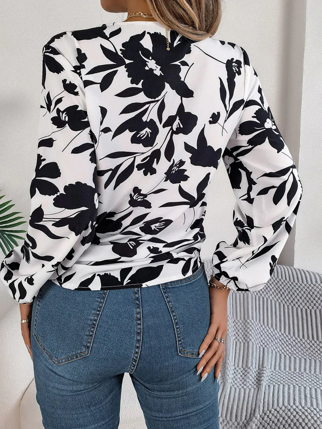 Back view of a woman wearing a white long-sleeve blouse with a black floral print and a tie-front detail, paired with blue denim jeans.
