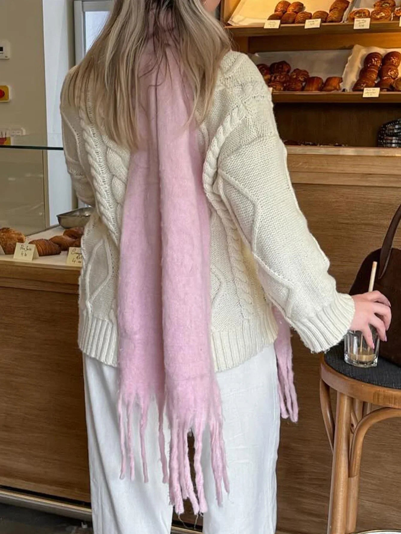 Back view of a woman wearing a cream cable knit sweater and white pants with a long pink scarf draped over her shoulders.