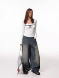 Full-body shot of a woman in a white ribbed long-sleeve top with a black bow accent, wearing wide-leg distressed denim pants and carrying a silver handbag.