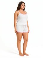 Woman wearing a white ribbed camisole and shorts pajama set with pink lace trim and bow details, side view, left.