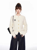 Woman wearing a cream cable knit cardigan with gold buttons and an embroidered heart detail on the left chest, paired with dark wash wide-leg jeans.