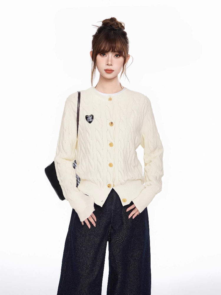 Woman wearing a cream cable knit cardigan with gold buttons and an embroidered heart detail on the left chest, paired with dark wash wide-leg jeans.