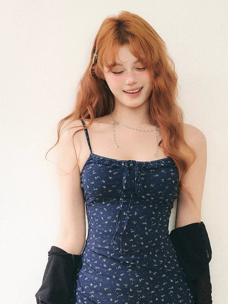 Woman wearing a navy floral print mini dress with spaghetti straps and a tie-front detail, paired with a sheer black cardigan and brown boots.
