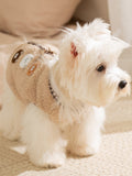 A white dog stands wearing a tan fleece vest featuring bear appliques and a buckle closure.
