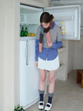 Woman examining items in an open refrigerator, dressed in a grey sweater, blue gingham shirt, and white skirt.