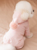 Rear view of a white Bichon Frise dog wearing a pink gingham hooded jumpsuit with a pink pom-pom, sitting on a bed