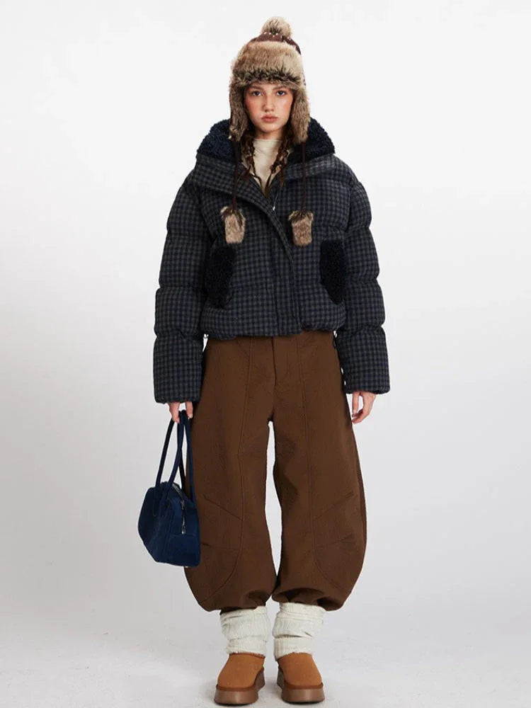 Woman wearing a cropped plaid puffer jacket with a fur-lined collar and hood, brown wide-leg pants, and a knit beanie with ear flaps.