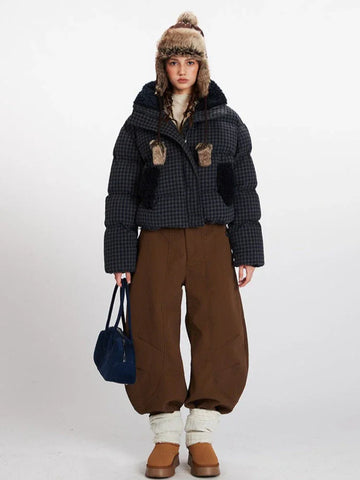 Woman wearing a cropped plaid puffer jacket with a fur-lined collar and hood, brown wide-leg pants, and a knit beanie with ear flaps.