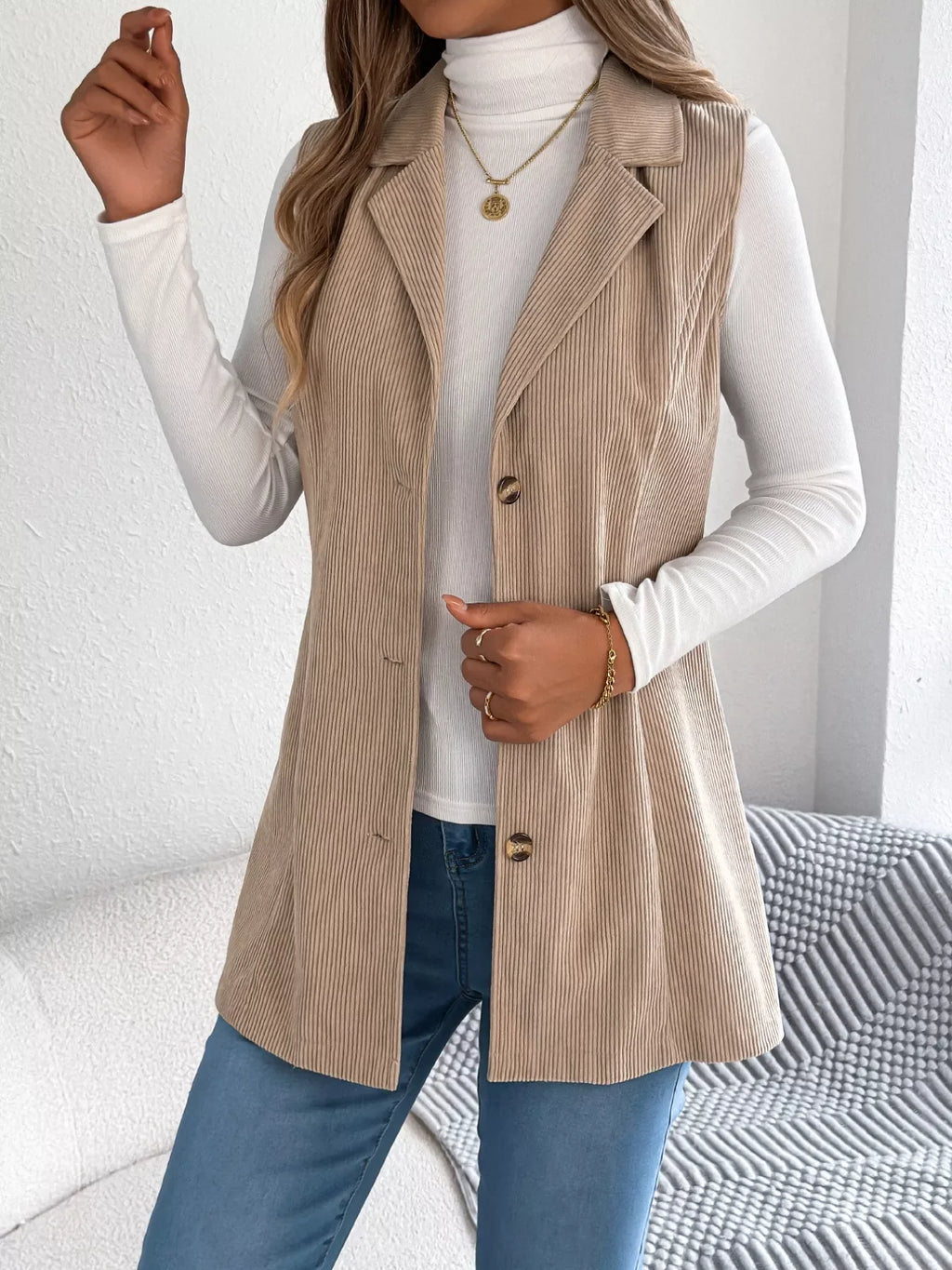 Woman posing in a beige corduroy longline vest, white turtleneck, and blue jeans, holding a white clutch.