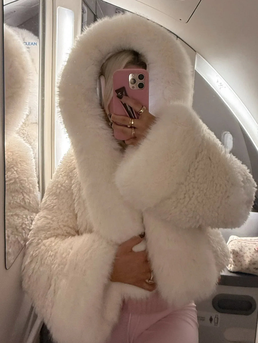 Woman taking a mirror selfie on an airplane wearing a cream-colored faux fur cropped jacket with an oversized hood