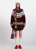 Woman wearing a burgundy Fair Isle crewneck sweater with an apple motif and a white knit hat