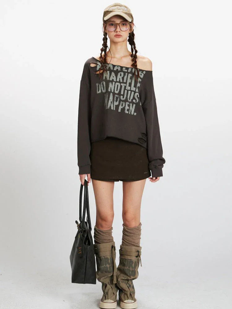 Woman wearing a dark grey distressed graphic print cropped sweatshirt with an off-the-shoulder neckline, paired with a dark brown mini skirt and knee-high socks with boots.