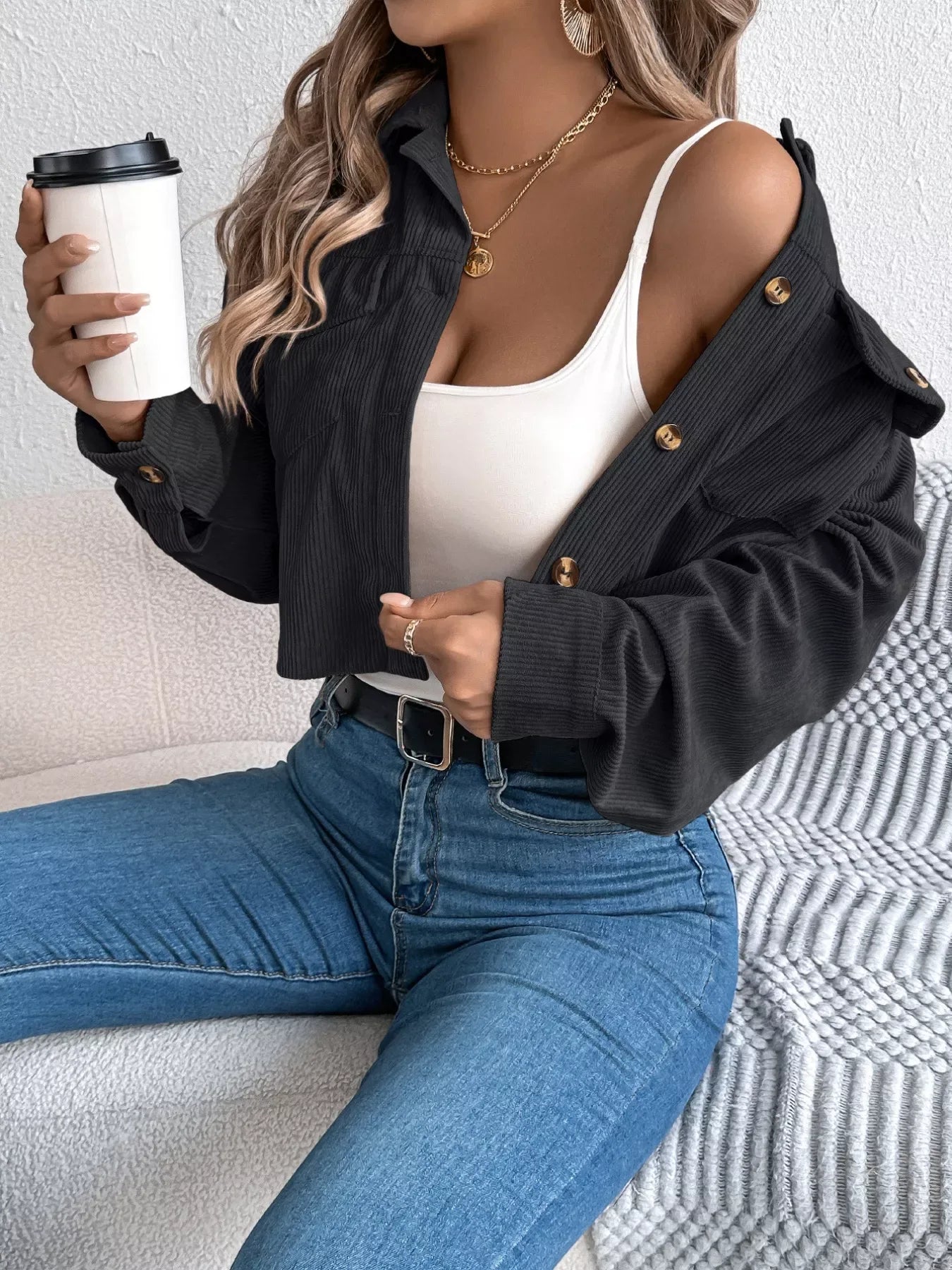 Woman holding a coffee cup while wearing a black cropped corduroy jacket and white tank top