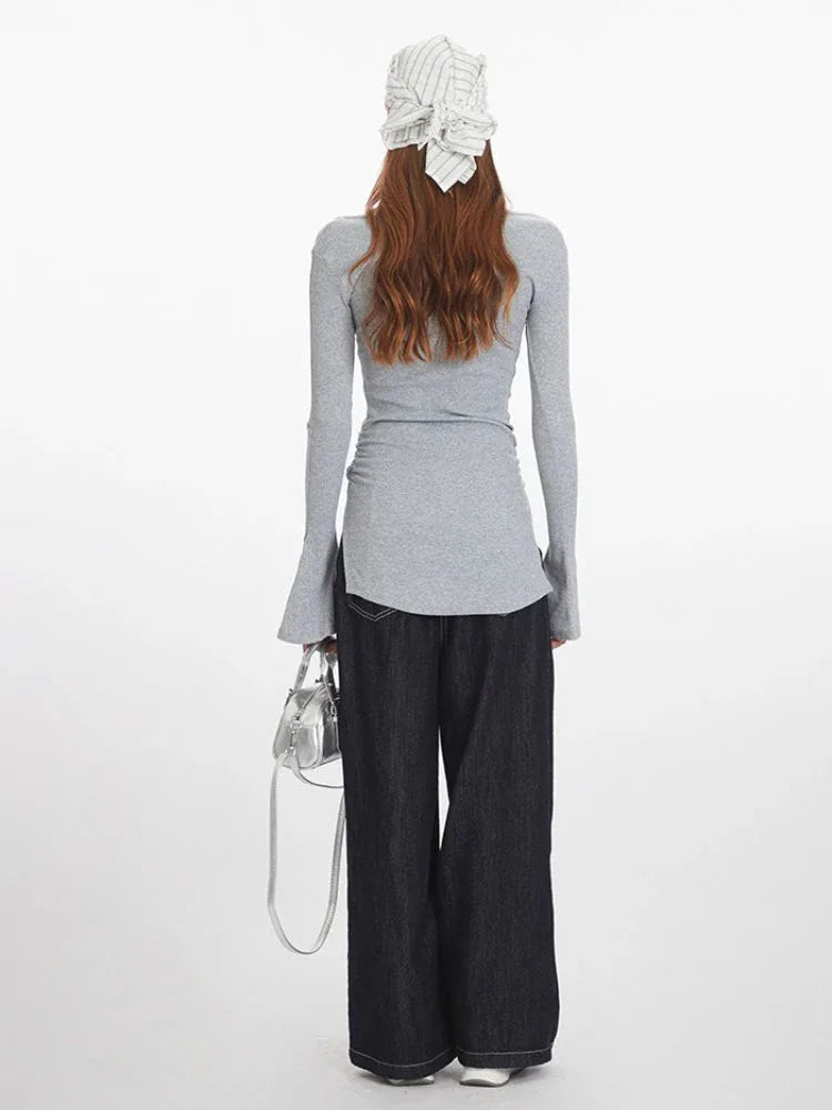 Woman wearing a grey long-sleeved ribbed knit top with a scoop neckline, ruched side detail, flared cuffs, and a side slit hem, paired with dark wash wide-leg jeans and a silver handbag.