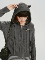 Model adjusting the hood of a grey cable knit cropped hooded jacket with ear details.