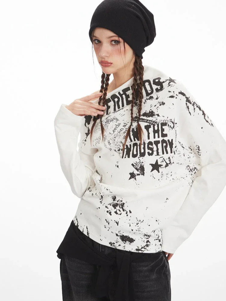 Front view of a woman wearing a white long-sleeve graphic print top with an off-the-shoulder design, styled with dark wash jeans and a black beanie.