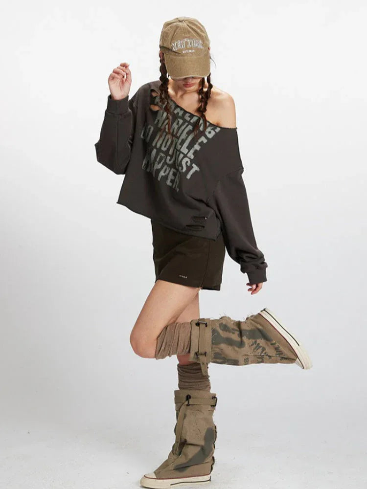 Woman posing in a dark grey distressed graphic print cropped sweatshirt with an off-the-shoulder neckline, dark brown mini skirt, and styled socks and boots.