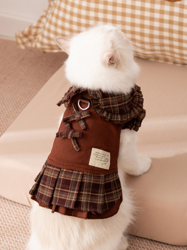 A white cat wearing a brown plaid pet dress with a ruffled collar and pleated skirt.