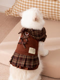 Back view of a white cat in a brown plaid pet dress with decorative bows and a D-ring leash attachment.